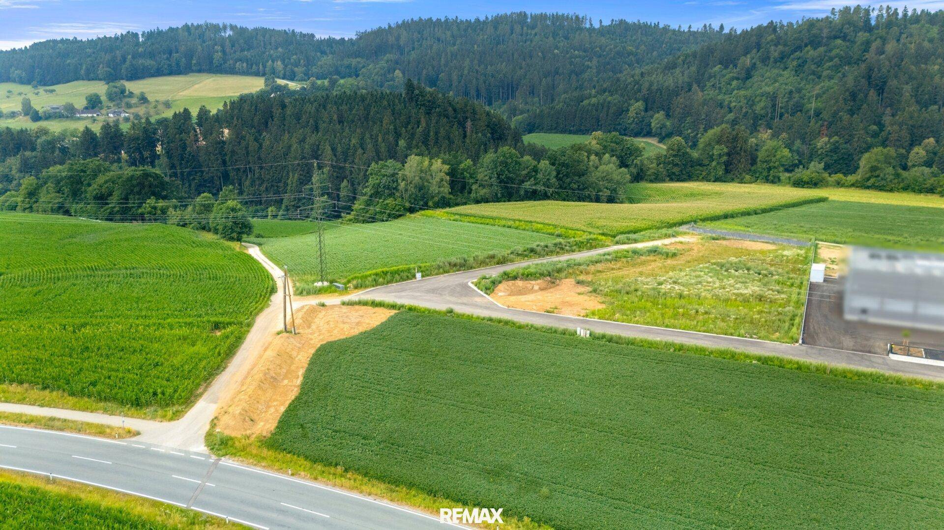 Weitläufige Landschaftsaufnahme mit grünen Feldern, Straßen und bewaldeten Hügeln im Hintergrund unter blauem Himmel.