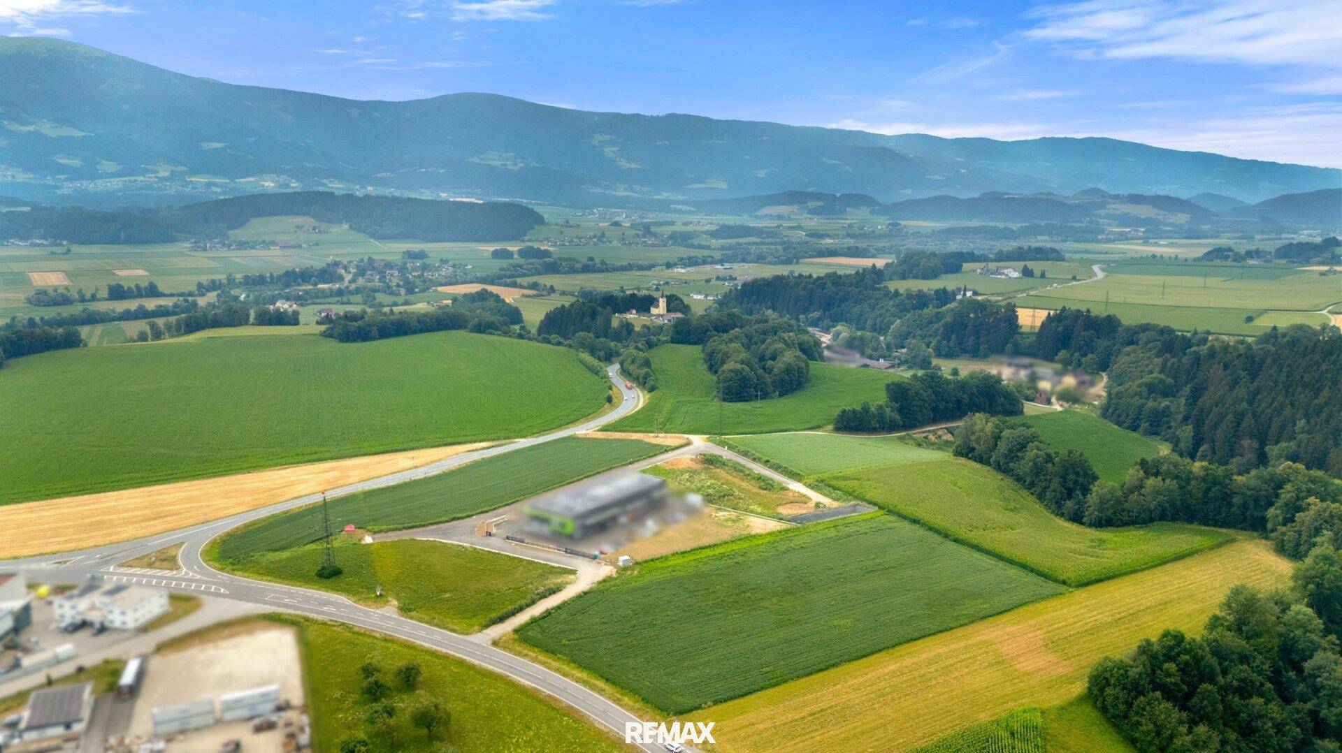 Panoramablick aus der Luft auf eine grüne Landschaft mit Feldern, Straßen und Bergen im Hintergrund.