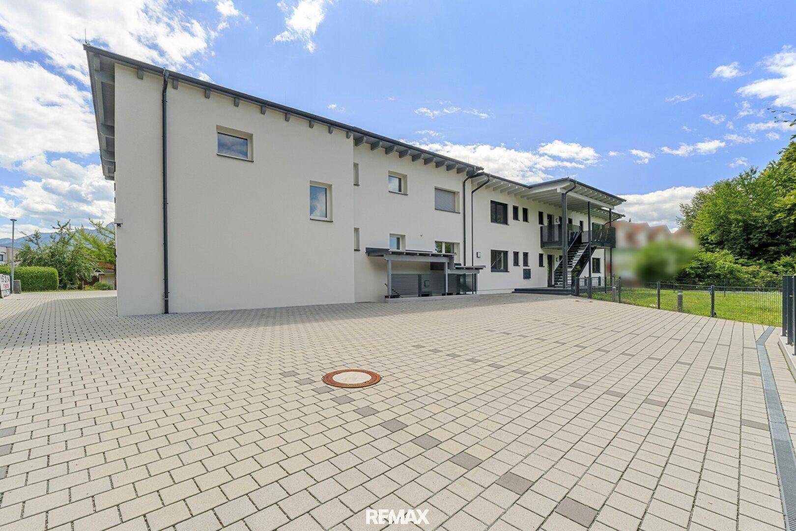 Seitenansicht des modernen Neubaus mit großem gepflasterten Parkplatz und Flachdach.