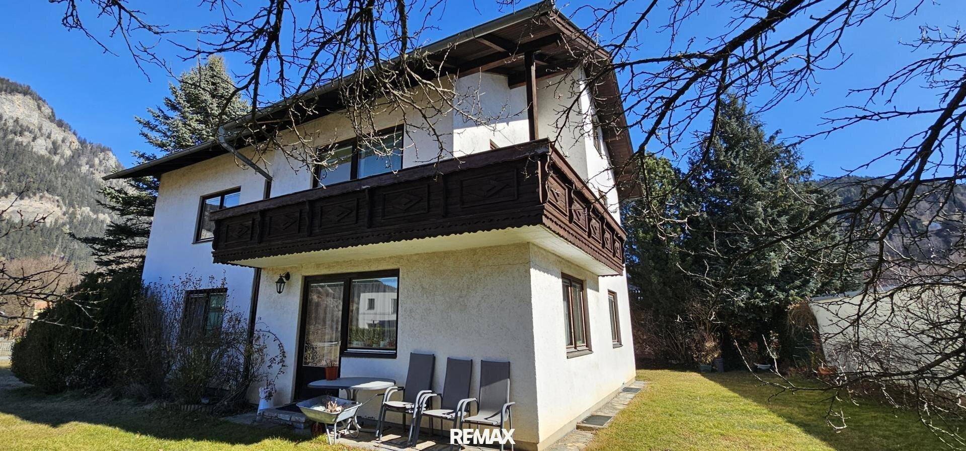 Gemütliche Terrasse mit Gartenmöbeln und Balkon darüber, umgeben von Natur und Bergen im Hintergrund.