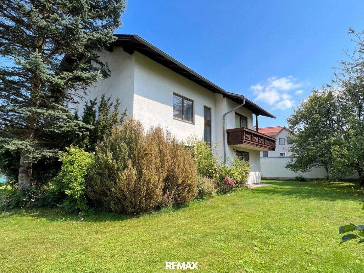 Seitenansicht des Hauses mit gepflegtem Garten, Büschen und einem Balkon unter blauem Himmel.