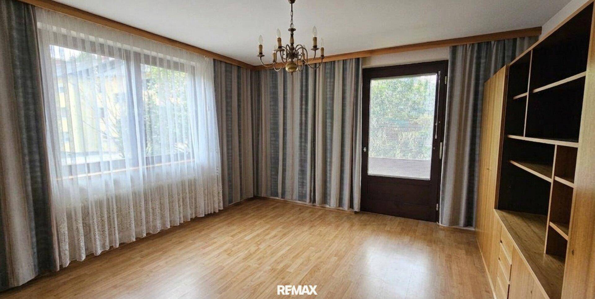 Geräumiges Wohnzimmer mit Holzboden, großen Fenstern und Zugang zu einem Außenbereich.