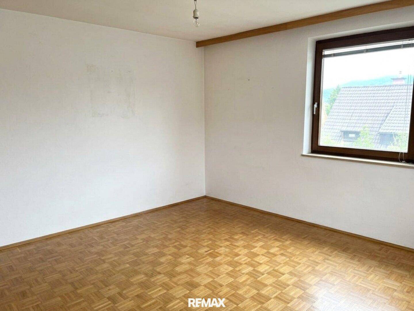Leeres Schlafzimmer mit Parkettboden, weiß gestrichenen Wänden und Fenster mit Ausblick ins Grüne.