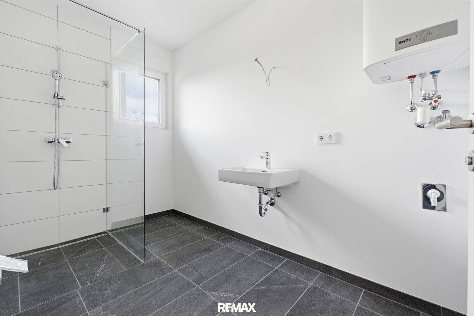 Modernes Badezimmer mit ebenerdiger Dusche, Wandwaschbecken und dunklen Fliesen, unmöbliert.