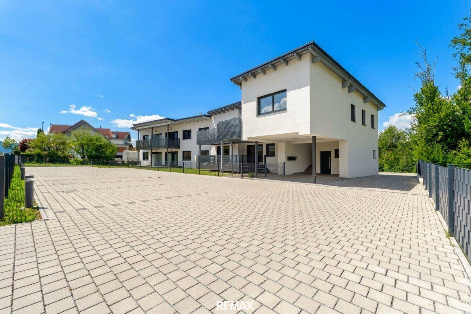 Neubau mit weißer Fassade und großem gepflasterten Parkplatz unter blauem Himmel.