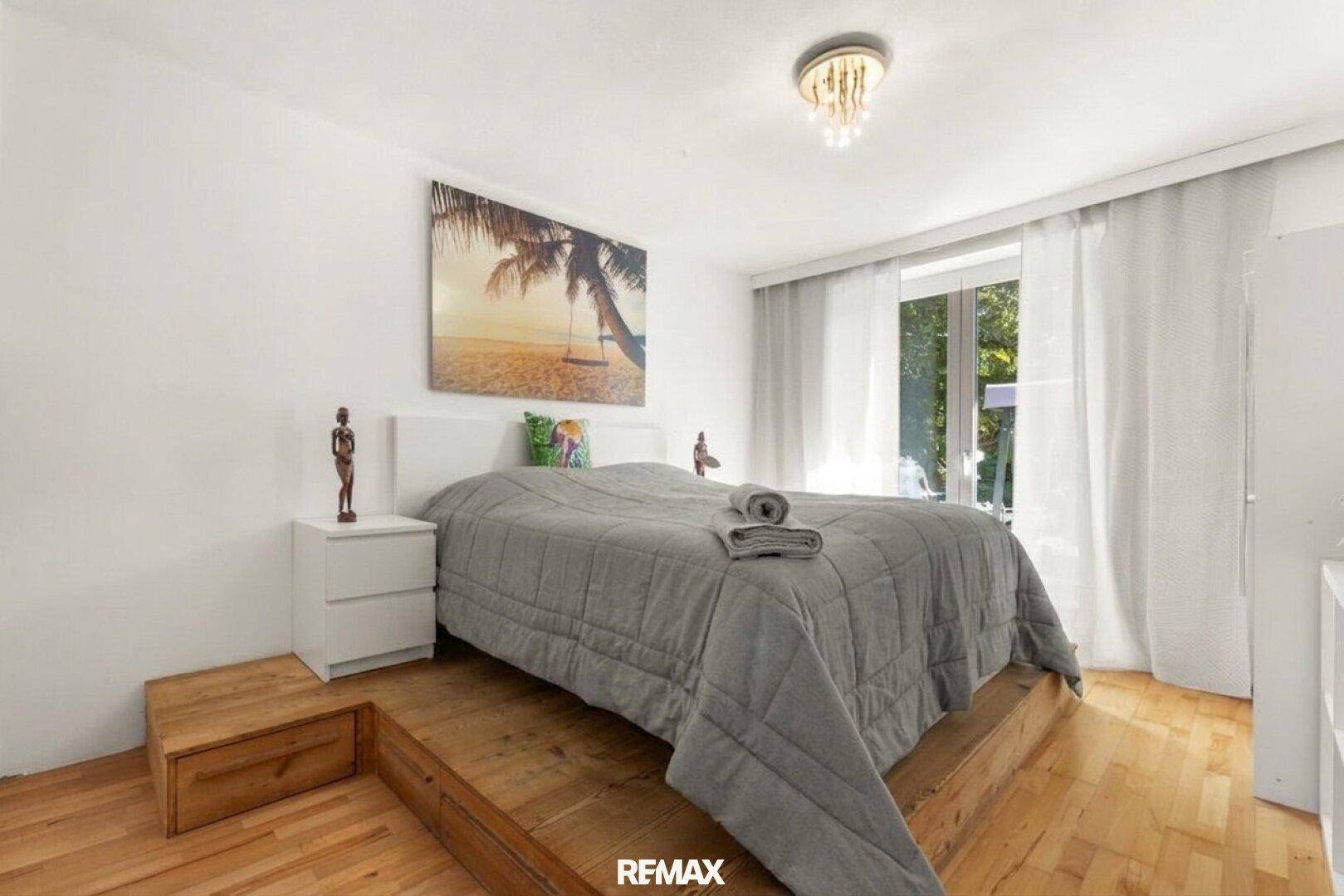 Modernes Schlafzimmer mit Holzbettgestell, weißer Einrichtung und großen Fenstern.