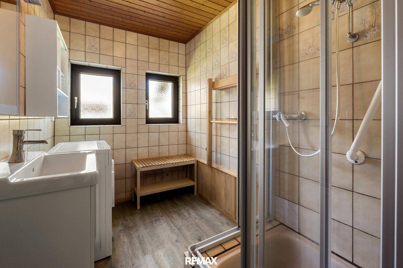 Gefliestes Badezimmer mit Dusche, Waschbecken und Fenster, ausgestattet mit Holzdetails.