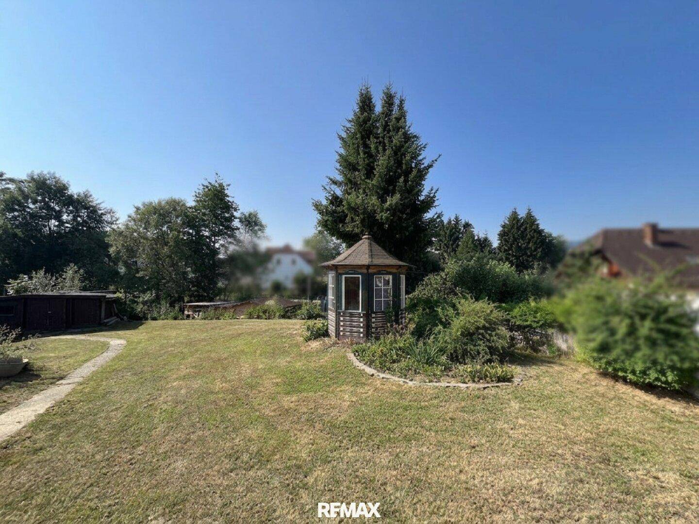 Großer Garten mit einem charmanten Holzpavillon und hohen Bäumen unter strahlend blauem Himmel.