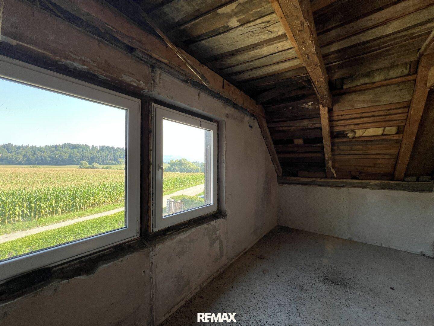 Dachzimmer mit freiliegenden Holzbalken, neuen Fenstern und weitem Blick über Felder.
