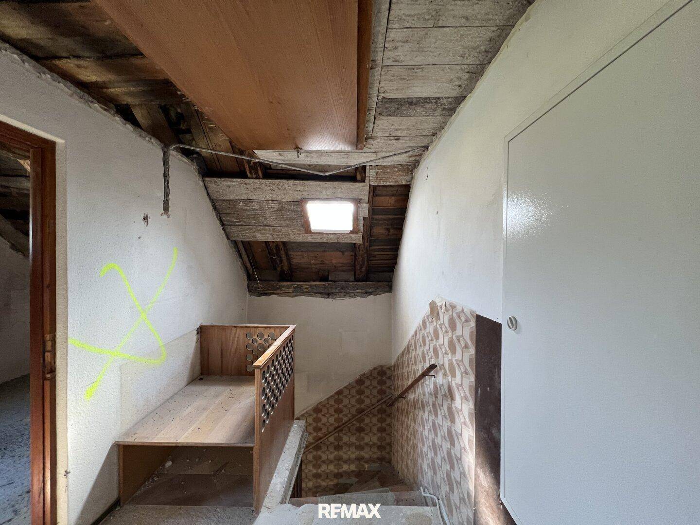 Treppenaufgang mit sichtbaren Holzbalken und einem Dachfenster, das Tageslicht spendet.