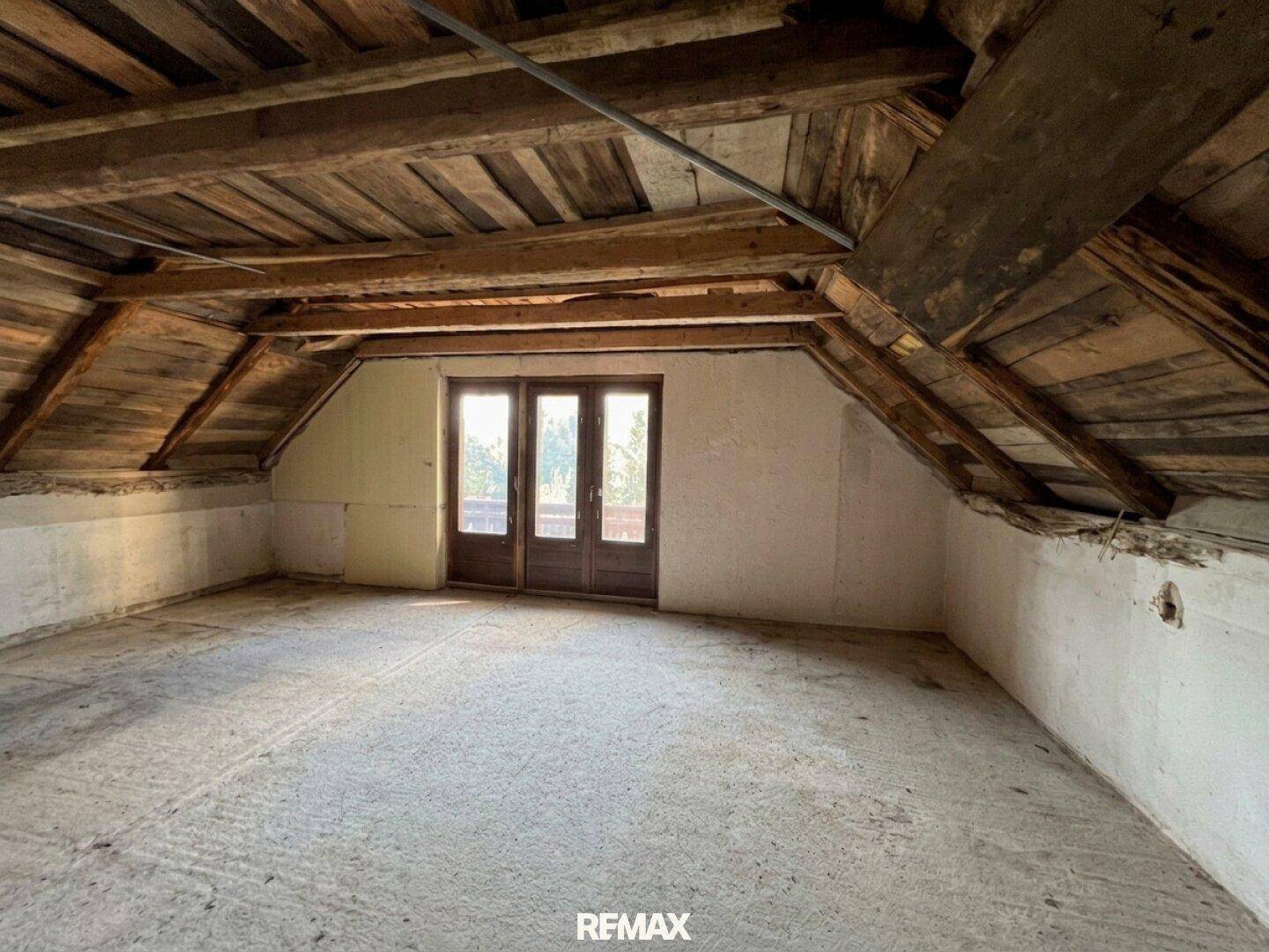 Geräumiges Dachzimmer mit sichtbaren Holzbalken und Doppeltüren, die ins Freie führen.