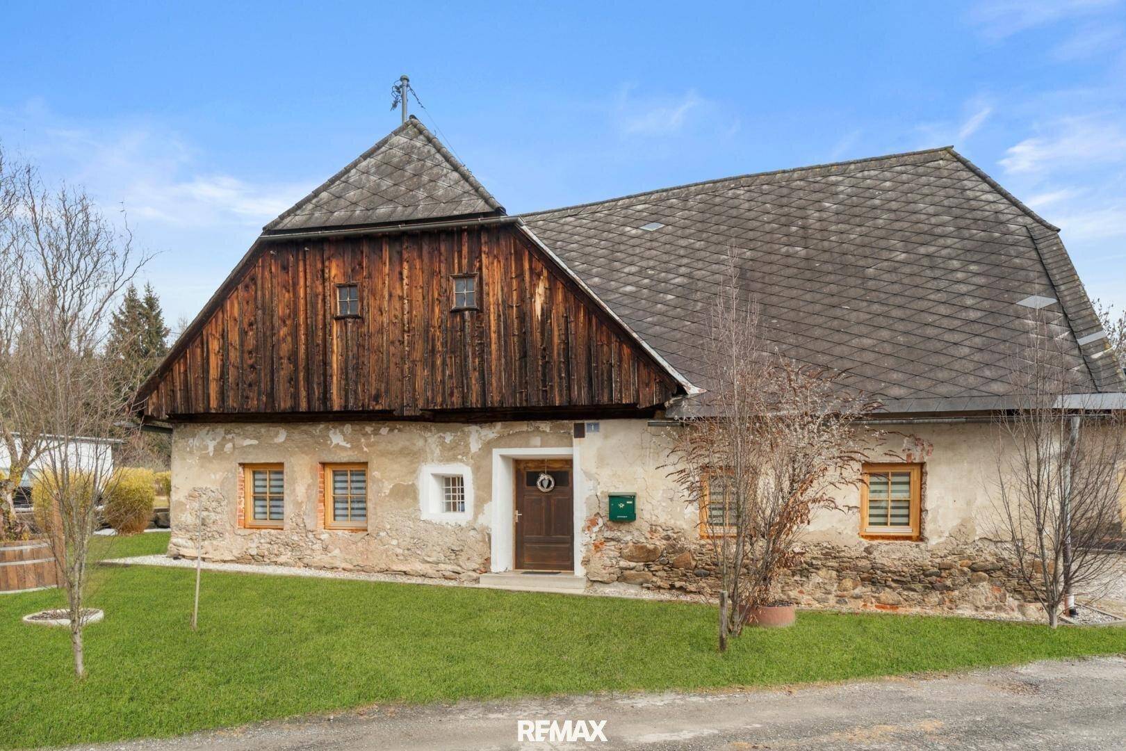 Traditionelles Bauernhaus mit Steinfassade, Holzanbau und Satteldach unter blauem Himmel.