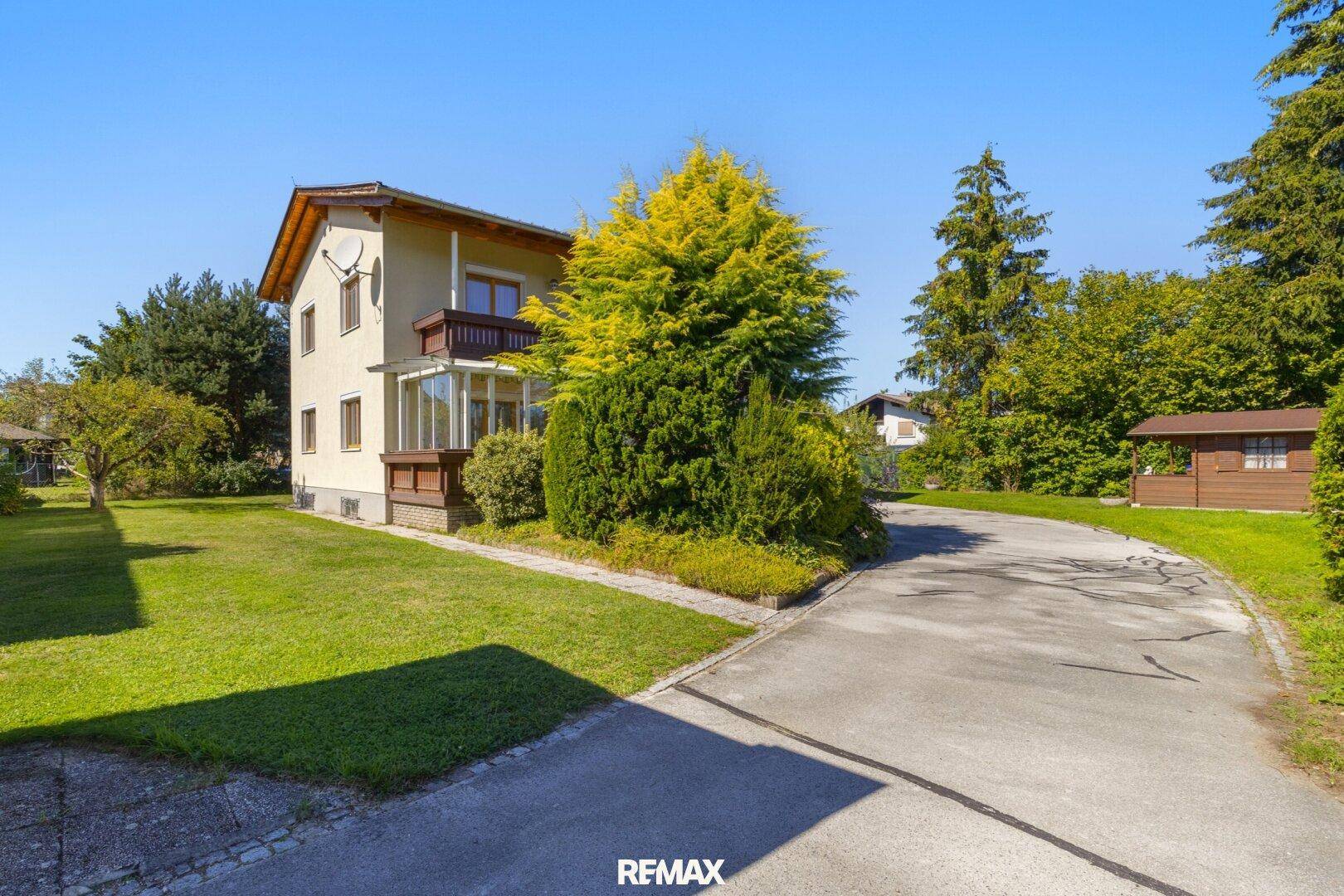 Das Haus von der Zufahrt aus gesehen, umgeben von gepflegtem Garten und Bäumen.