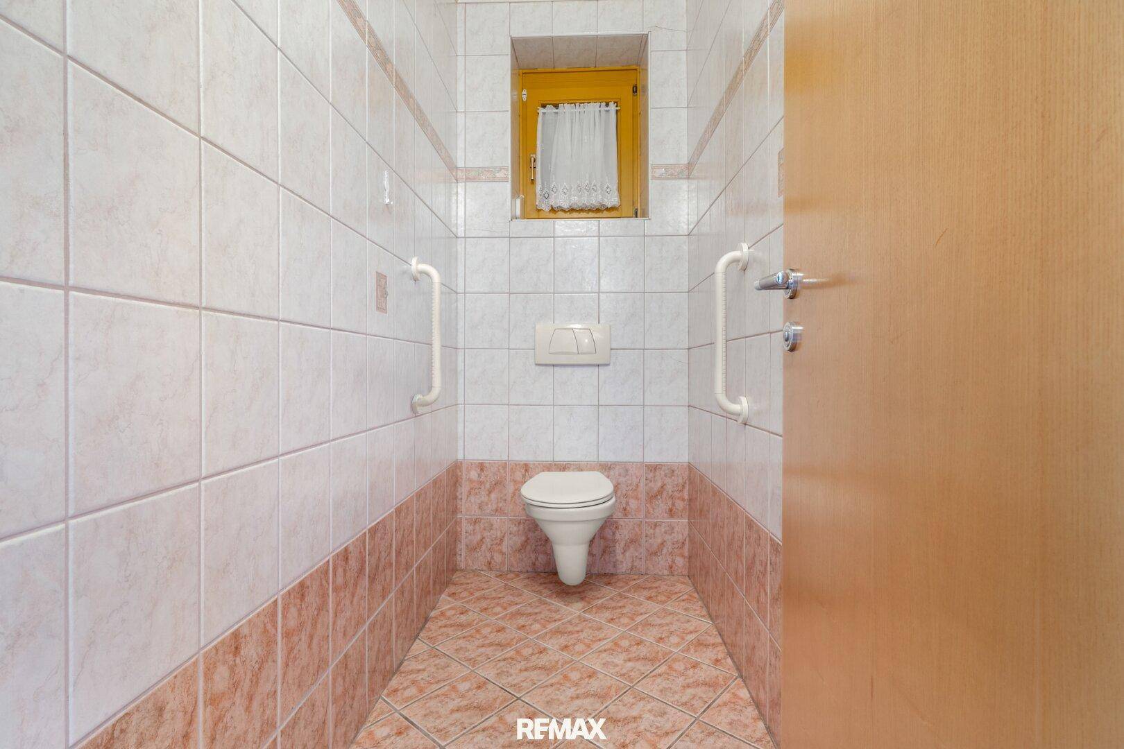 Separate Toilette mit beigefarbenen Fliesen und Haltegriffen für Barrierefreiheit.