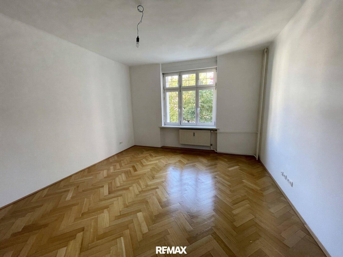 Geräumiges Zimmer mit klassischem Fischgrätparkett und einem großen Fenster für viel Helligkeit.