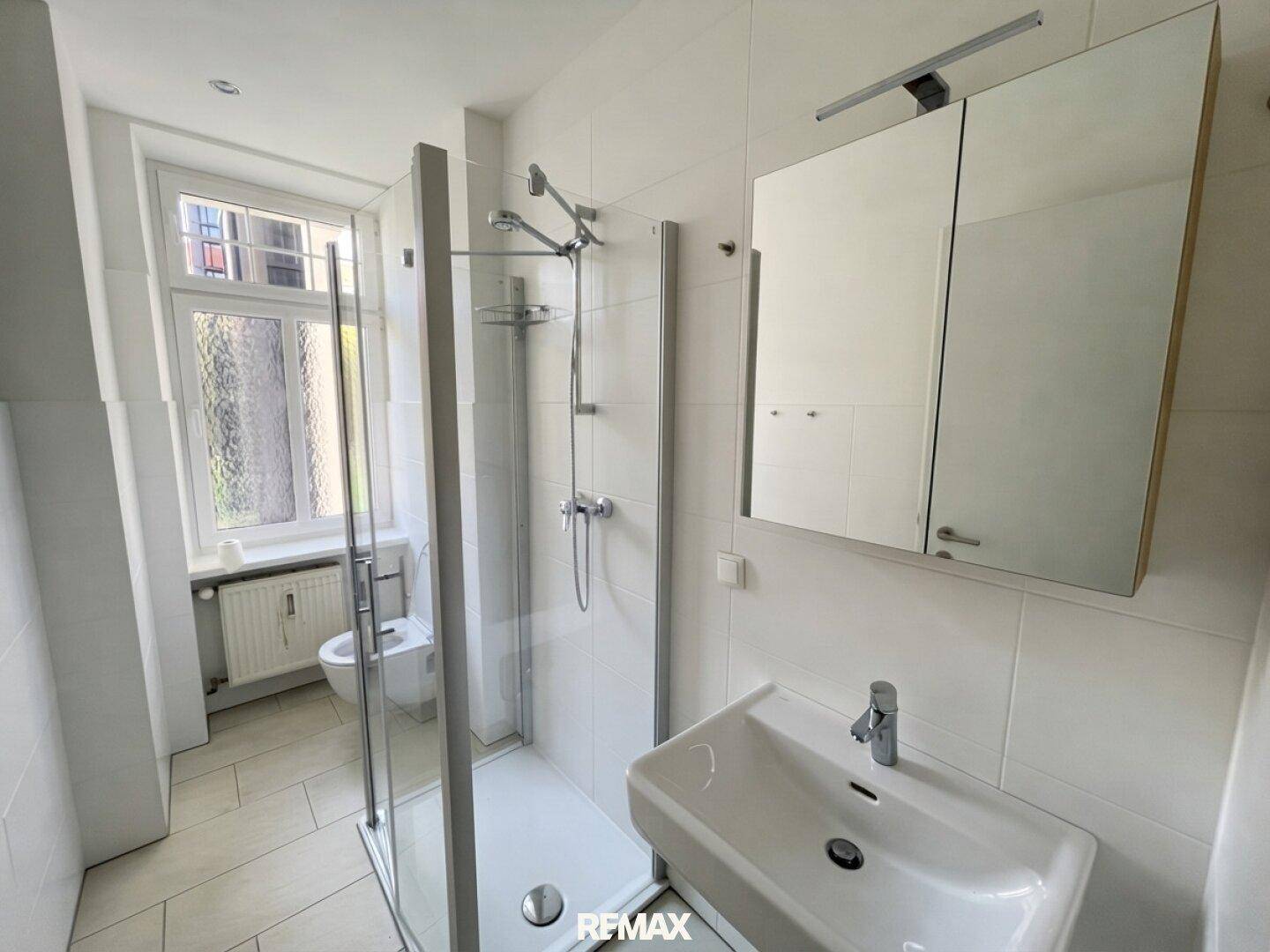 Modernes Badezimmer mit weißen Fliesen, begehbarer Dusche, Waschbecken und Spiegelschrank.