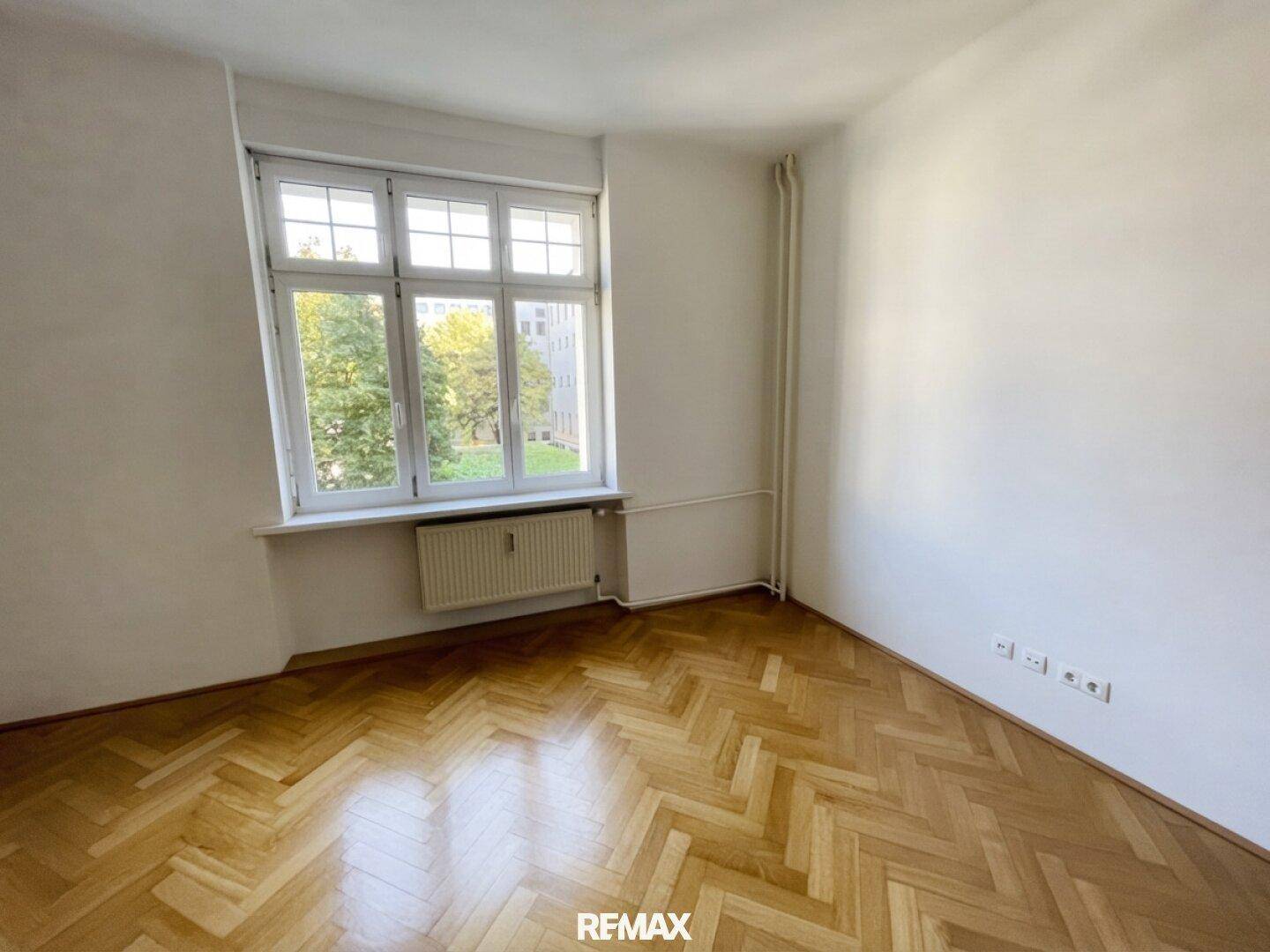 Helles Zimmer mit Fischgrätparkett, weiß gestrichenen Wänden und einem großen Fenster mit Ausblick.