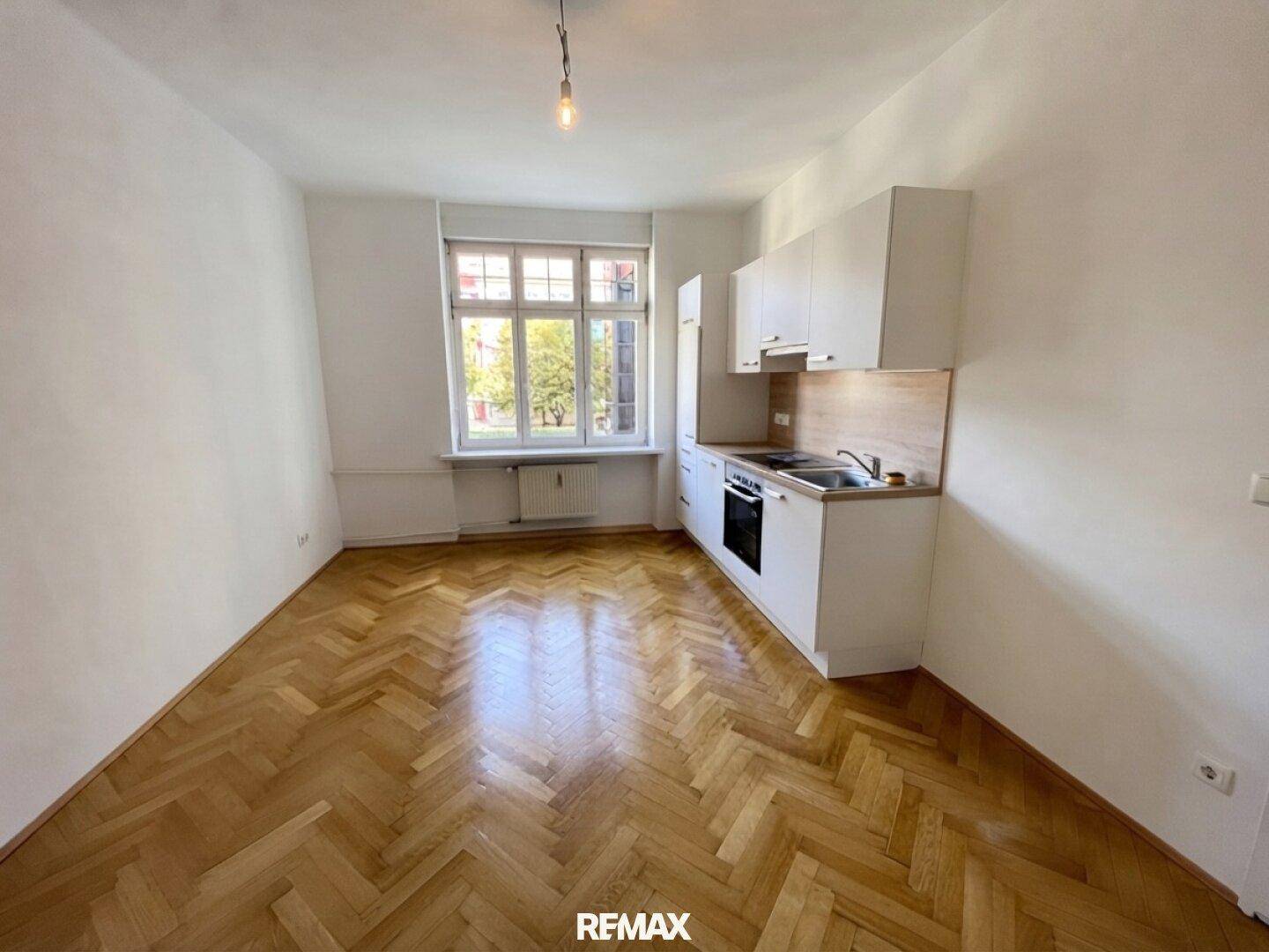 Heller Raum mit integrierter Küchenzeile, Fischgrätparkett und Blick ins Grüne durch das Fenster.