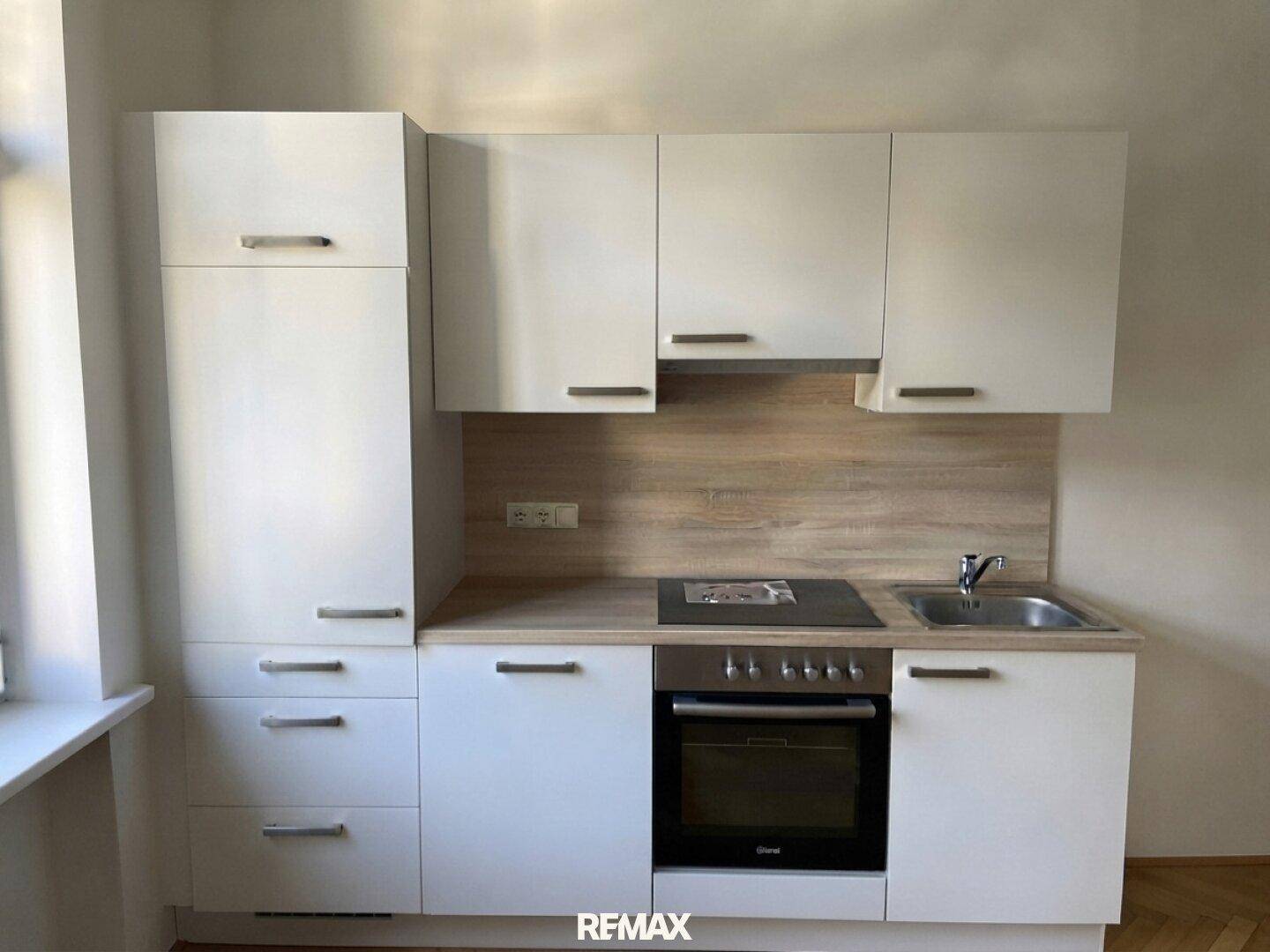 Moderne Einbauküche mit weißen Schränken, Holzoptik-Rückwand und integrierten Geräten, gut beleuchtet durch ein Fenster.