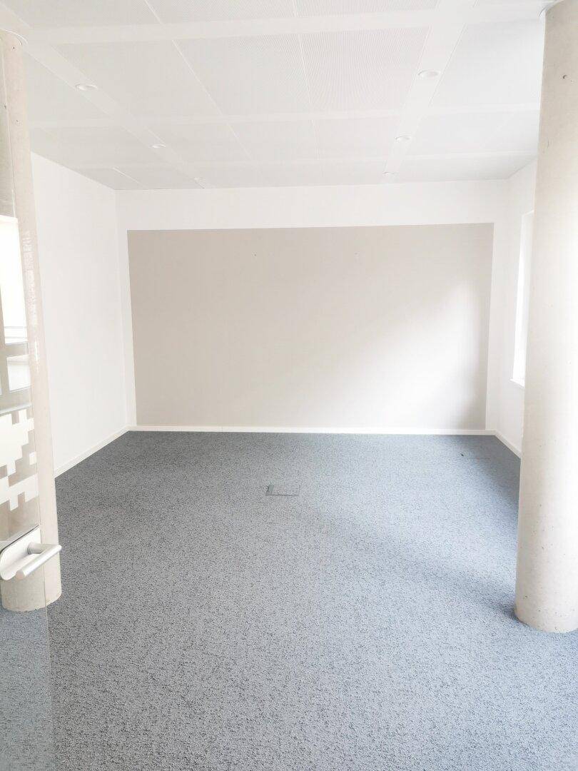 Heller Büroraum mit grauem Teppichboden, weißen Wänden und einer beigen Akzentwand, inklusive Betonsäule.
