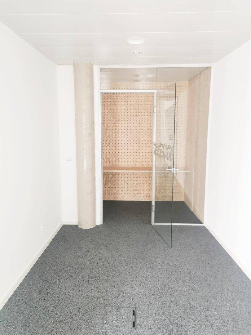 Leerer Büroraum mit grauem Teppichboden und einer gläsernen Trennwand zu einem kleinen Arbeitsbereich.