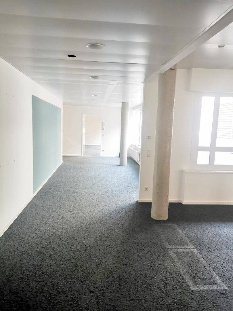 Langer, leerer Bürogang oder offener Arbeitsbereich mit grauem Teppichboden und Betonsäulen.