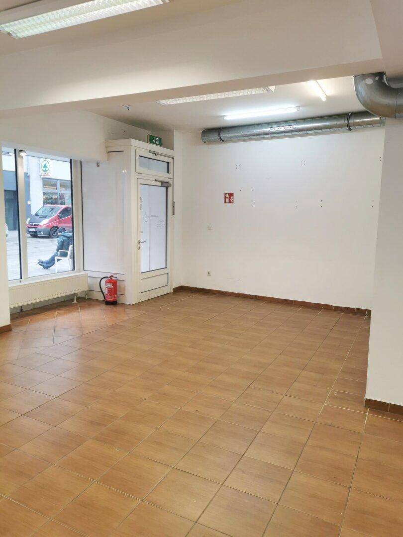 Leerer Ladenraum mit Fliesenboden, großen Schaufenstern und einem Aufzug im Hintergrund.