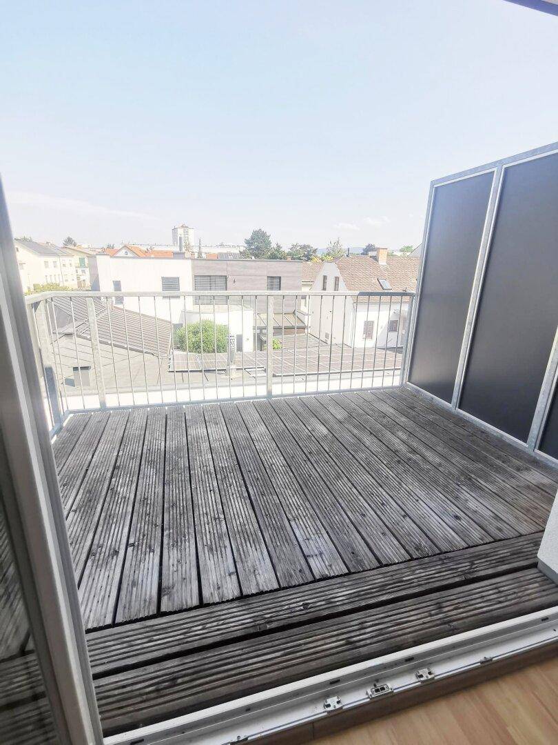 Balkon mit Holzbelag und Blick über die Dächer der Stadt, sonnig und offen.
