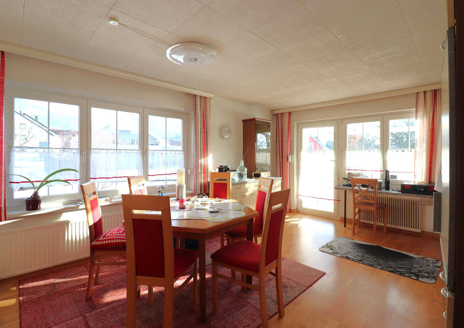 Gemütlicher Essbereich mit Holztisch, roten Stühlen und viel Tageslicht durch große Fenster.
