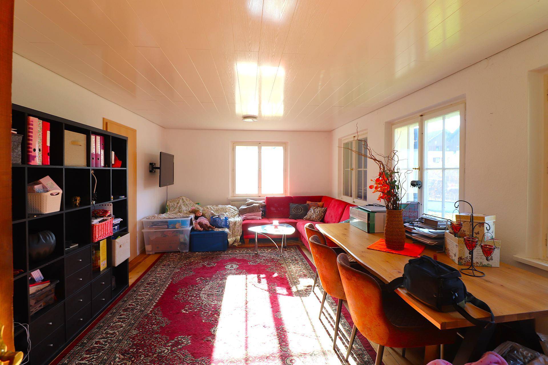 Geräumiges Wohnzimmer mit Teppichboden, rotem Sofa und viel Tageslicht durch große Fenster.