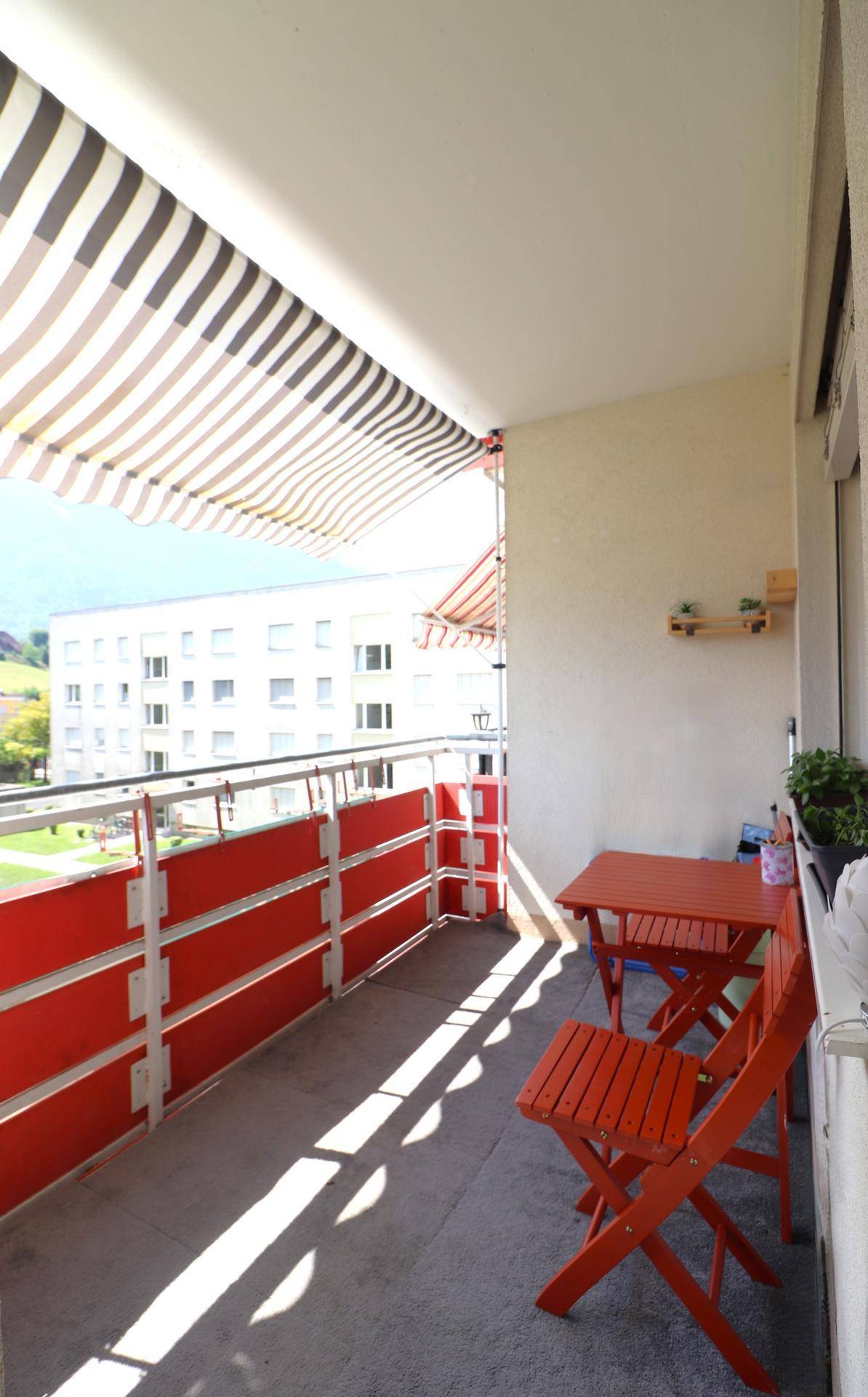 Sonniger Balkon mit Markise und roter Sitzgruppe, bietet Blick auf die umliegenden Gebäude.