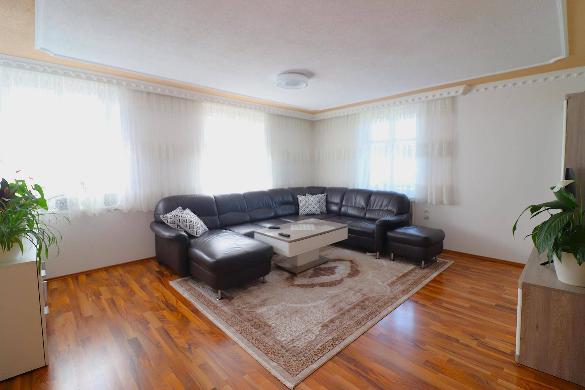 Gemütliches Wohnzimmer mit dunklem Ledersofa, Holzboden und hellen Vorhängen an den Fenstern.