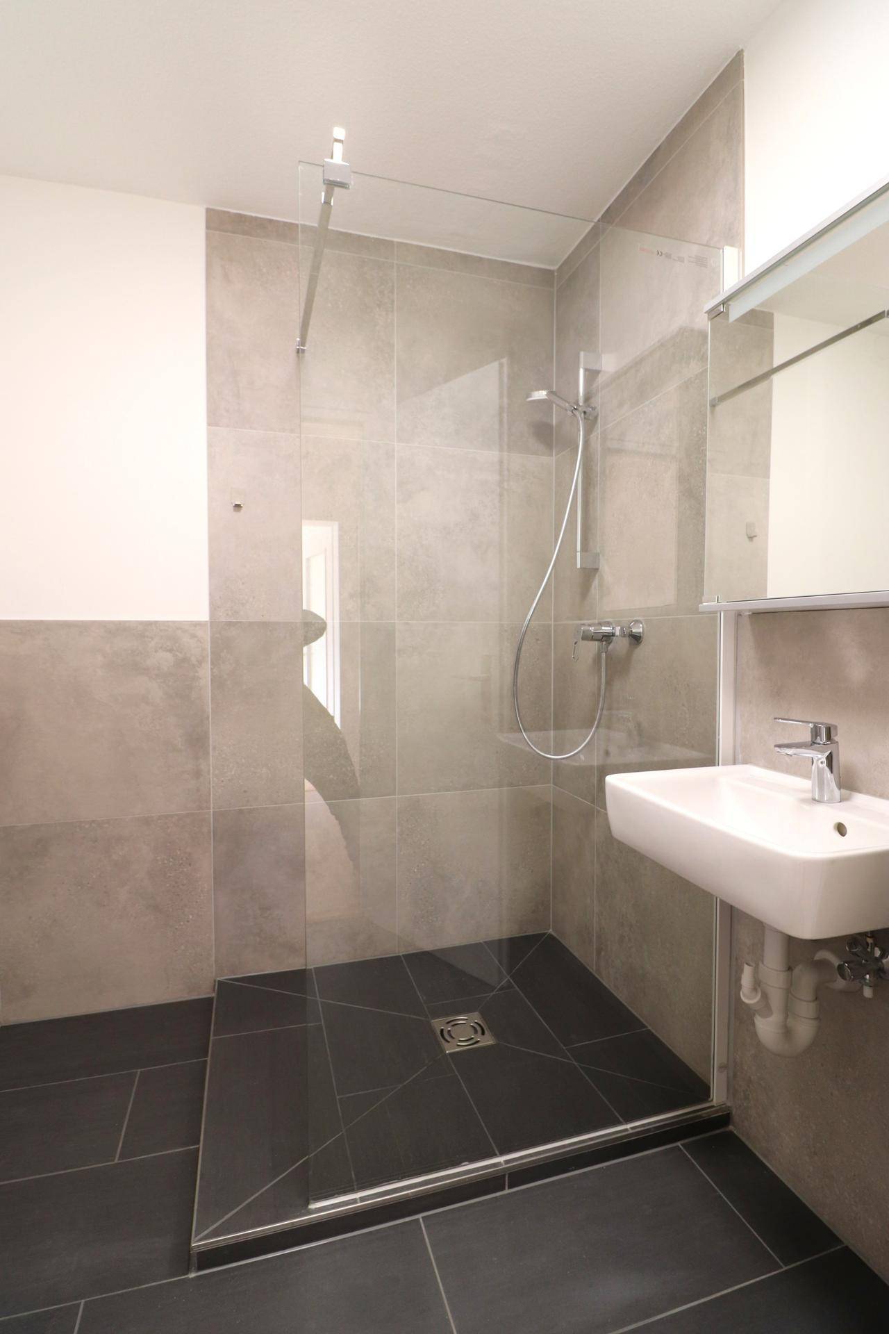 Modernes Badezimmer mit ebenerdiger Dusche, grauen Fliesen und kleinem Waschbecken.