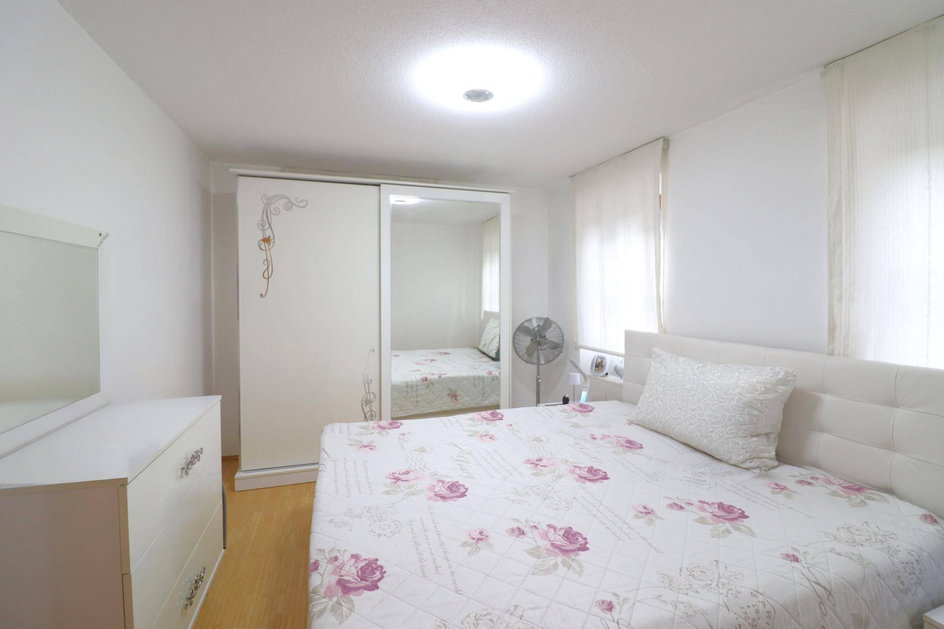Helles Schlafzimmer mit Doppelbett, Spiegelschrank und floraler Bettwäsche, gut beleuchtet.