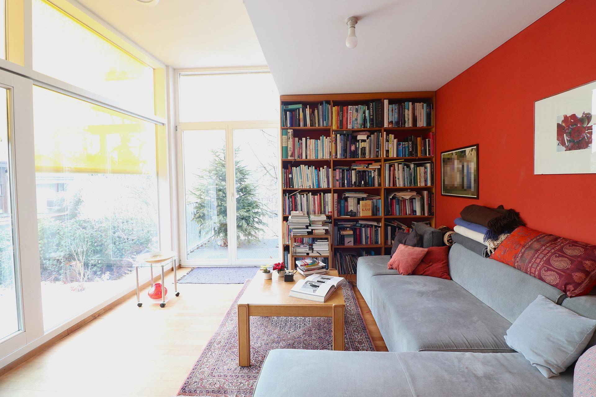 Helles Wohnzimmer mit roter Akzentwand, Bücherregal und Zugang zum Gartenbereich.