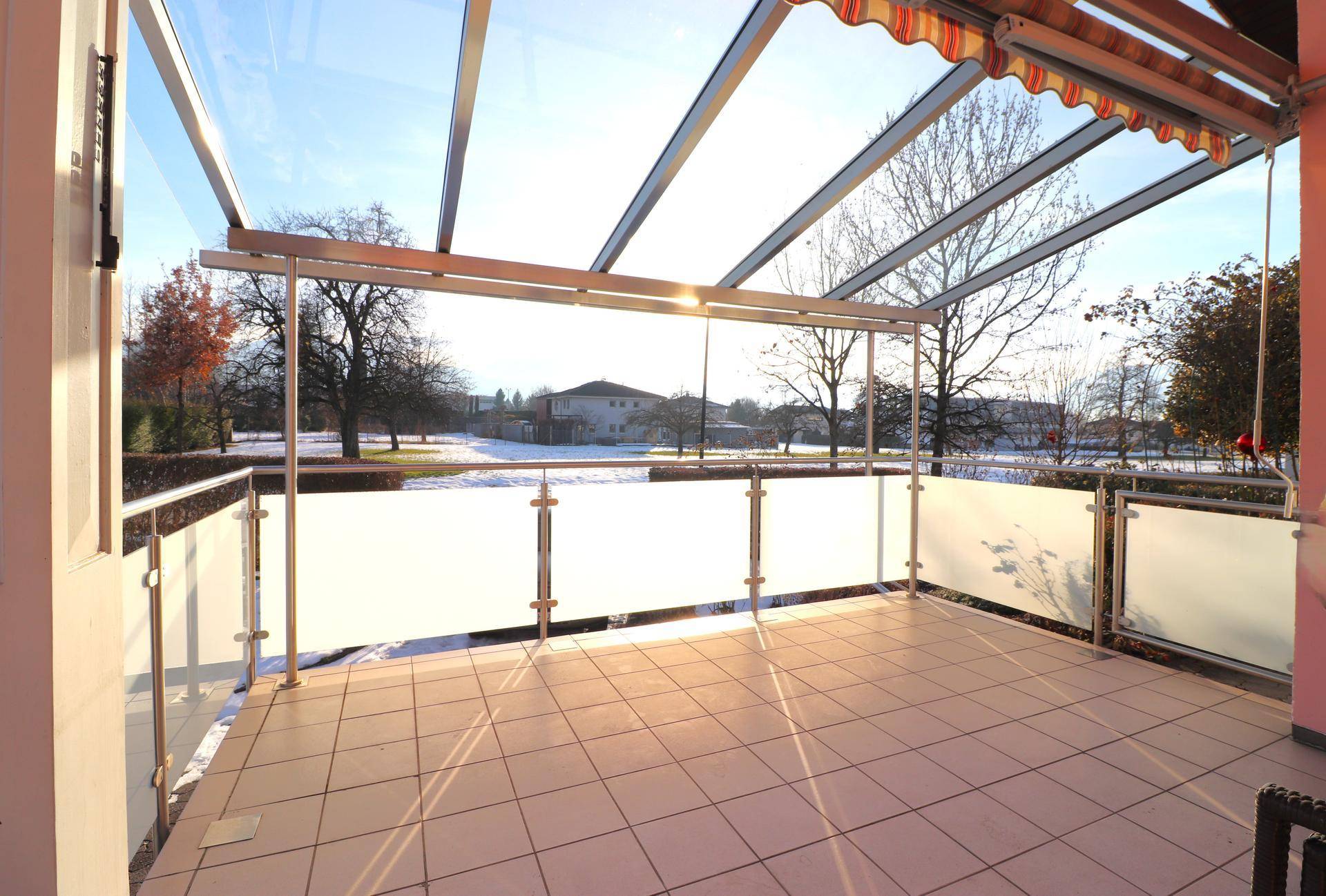 Überdachte Terrasse mit Fliesenboden und Glasgeländer, bietet einen weiten Blick ins Grüne.