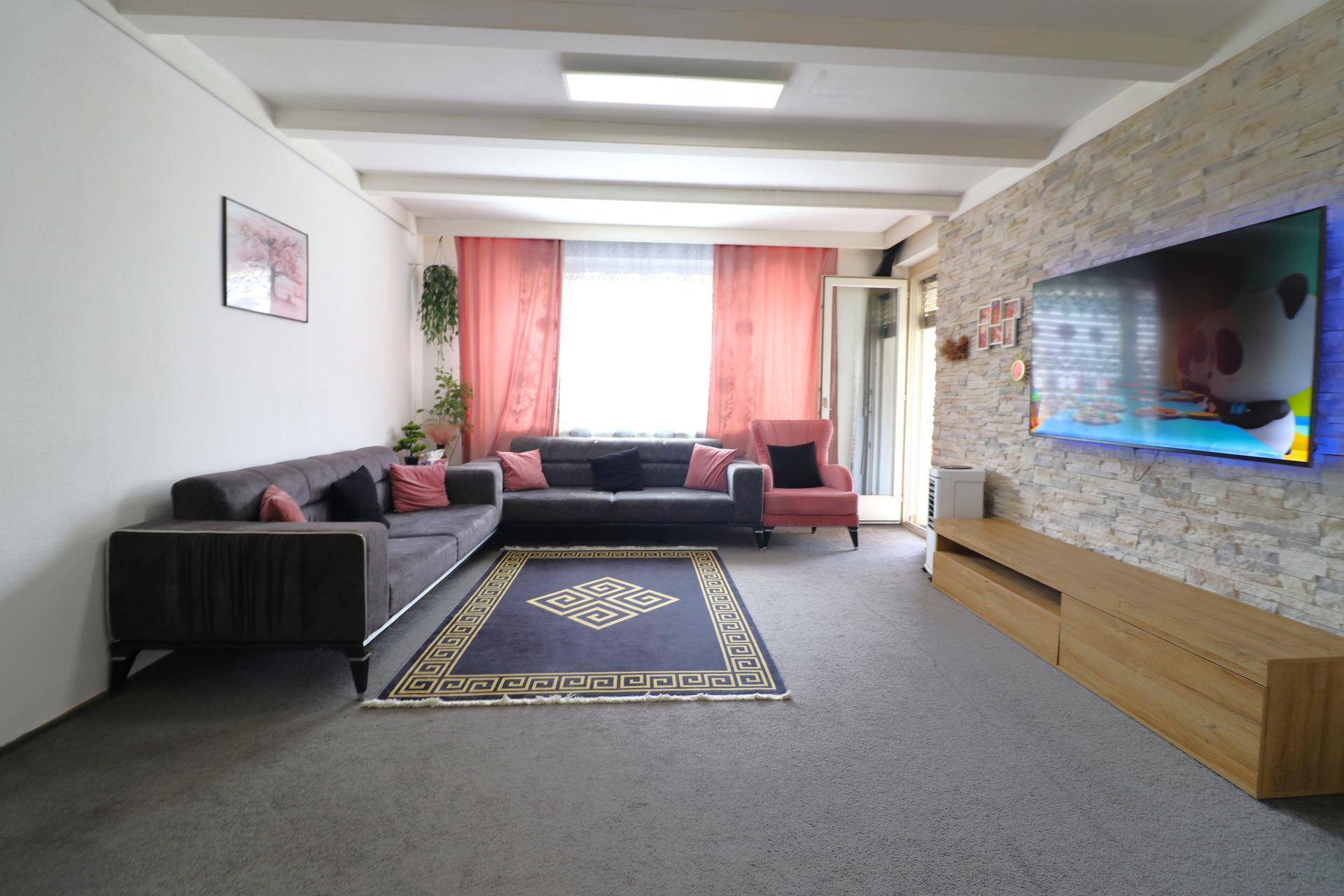 Helles Wohnzimmer mit Steinwand, Fernseher und Zugang zum Balkon, ausgestattet mit Teppichboden.