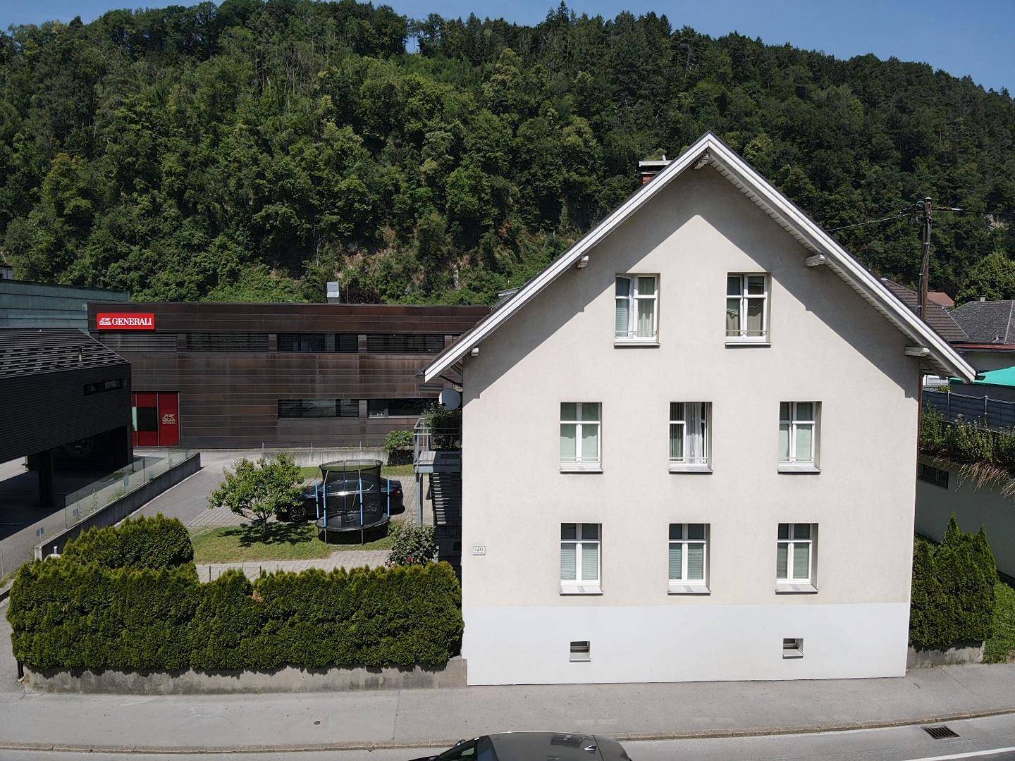 Vorderansicht des Hauses mit grüner Hecke, Parkplatz und bewaldetem Hügel im Hintergrund.