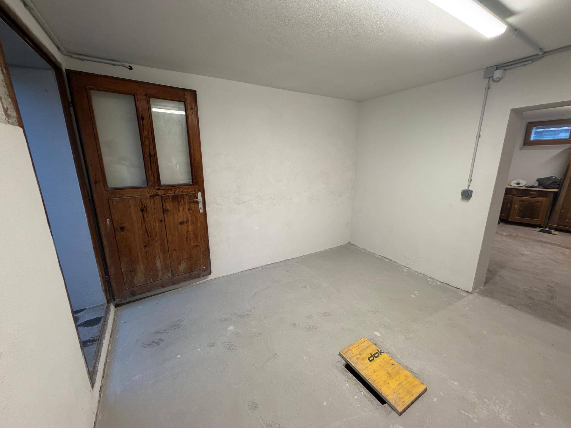 Ein weiterer leerer Kellerraum mit Betonboden, weiß gestrichenen Wänden und einer rustikalen Holztür.