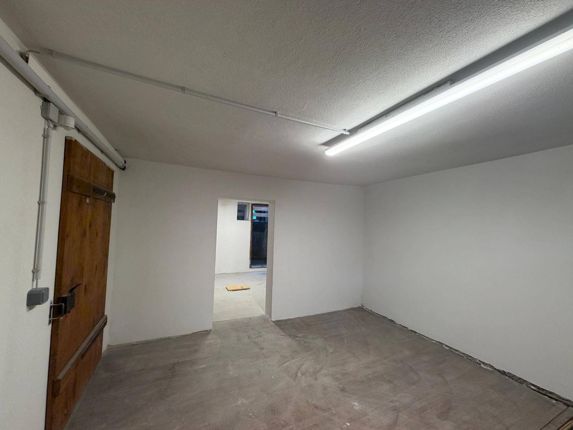 Ein leerer Kellerraum mit frisch gestrichenen weißen Wänden, Betonboden und sichtbaren Leitungen an der Decke.