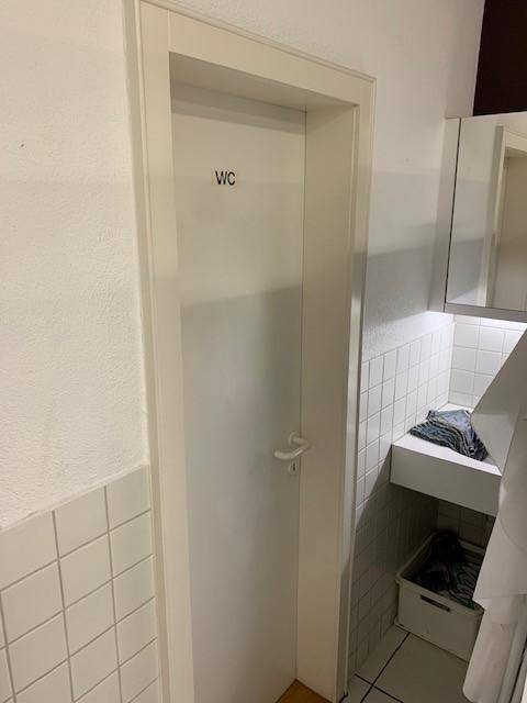 Weiß gestrichene Tür mit der Aufschrift "WC" neben einem gefliesten Bereich, der zu einer Toilette führt.