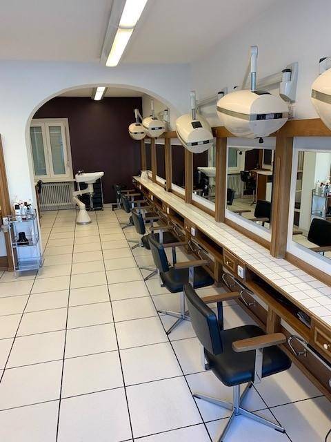 Langer Bereich im Friseursalon mit mehreren Frisierplätzen, Spiegeln, Trockenhauben und weißen Fliesenboden.