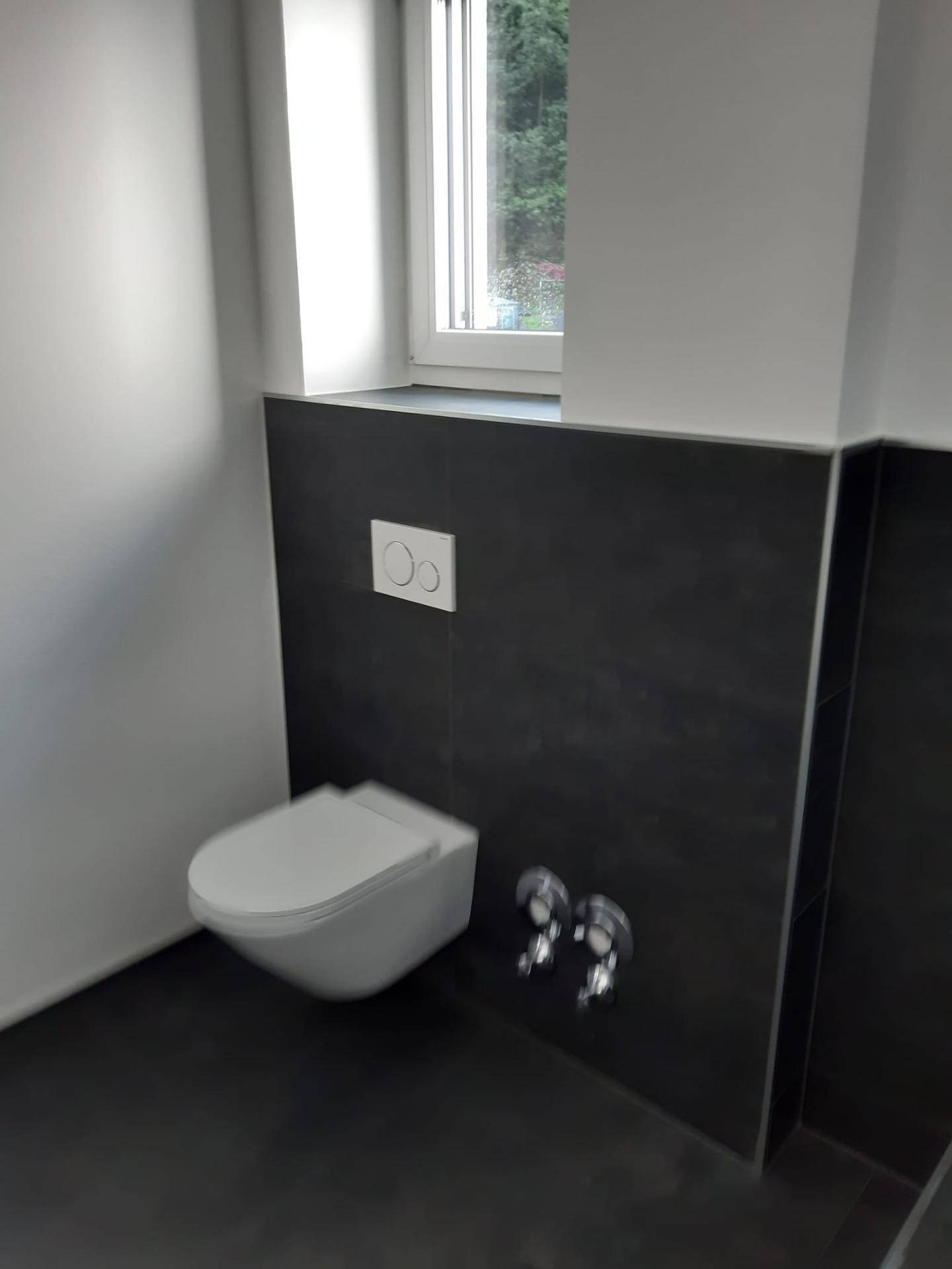 Wandhängende Toilette in einem Badezimmer mit dunklen Fliesen und einem Fenster für Tageslicht.