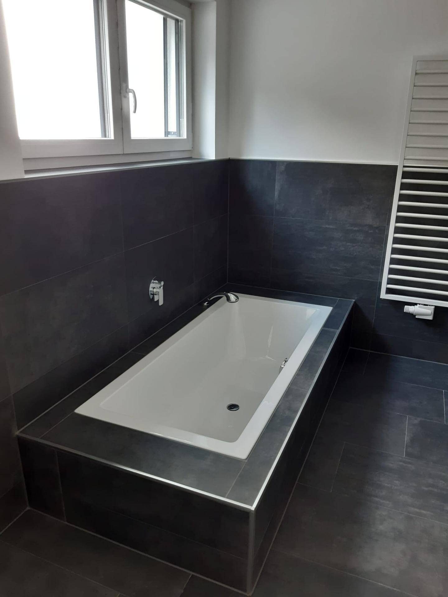 Eingebaute Badewanne mit dunklen Fliesen, einem Fenster und einem praktischen Handtuchheizkörper im Badezimmer.