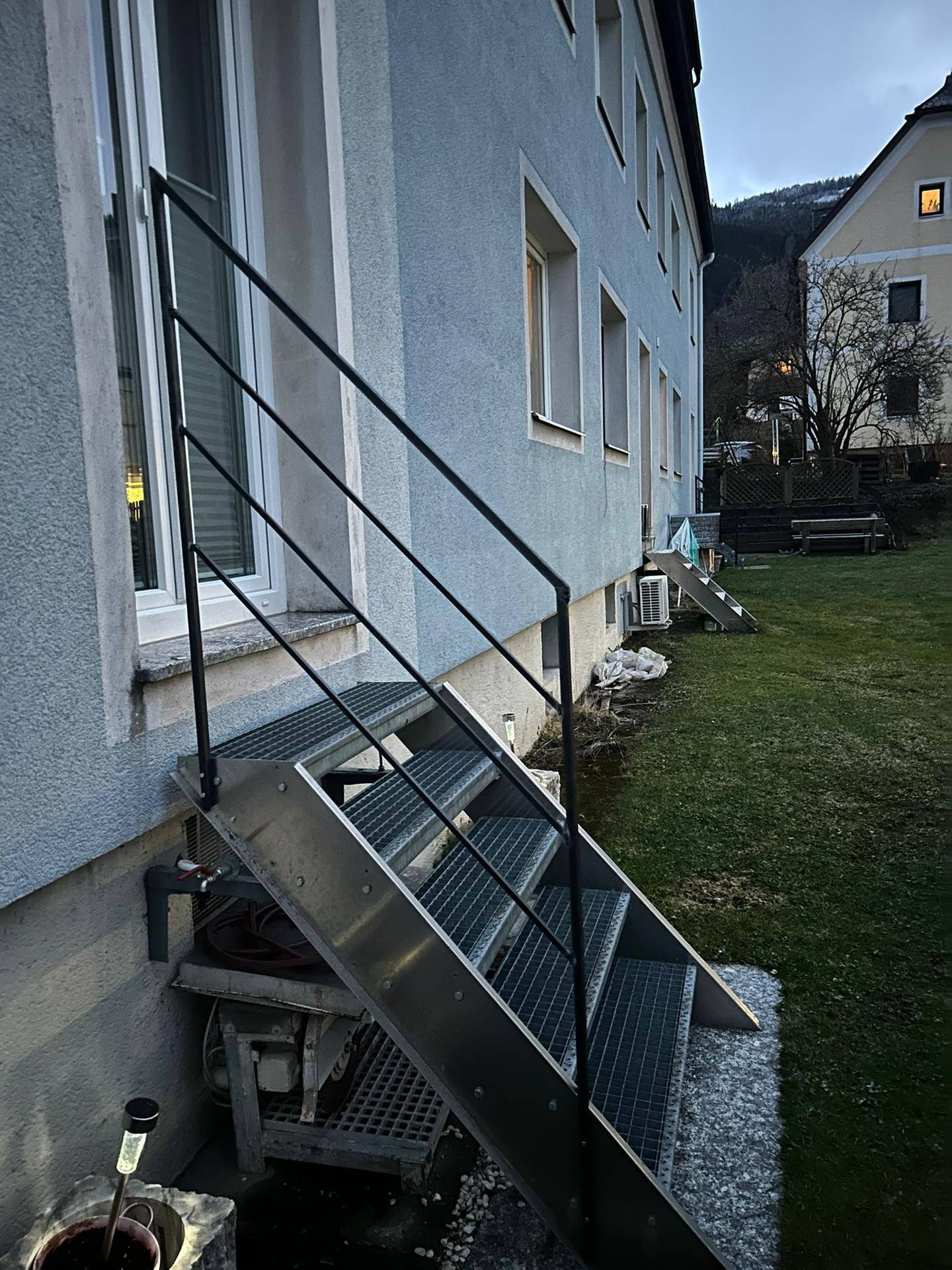 Hinterhofbereich des Gebäudes mit Metalltreppe, Rasenfläche und Blick auf die umliegende Landschaft.