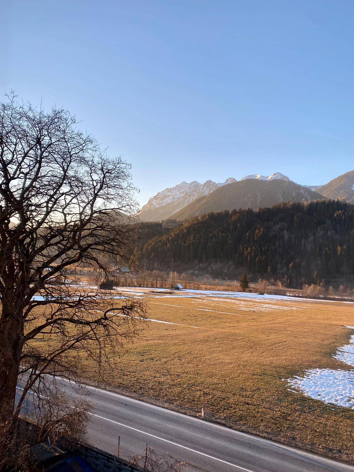 Sonnige Winterlandschaft mit schneebedeckten Feldern, Bergen und einem klaren blauen Himmel.