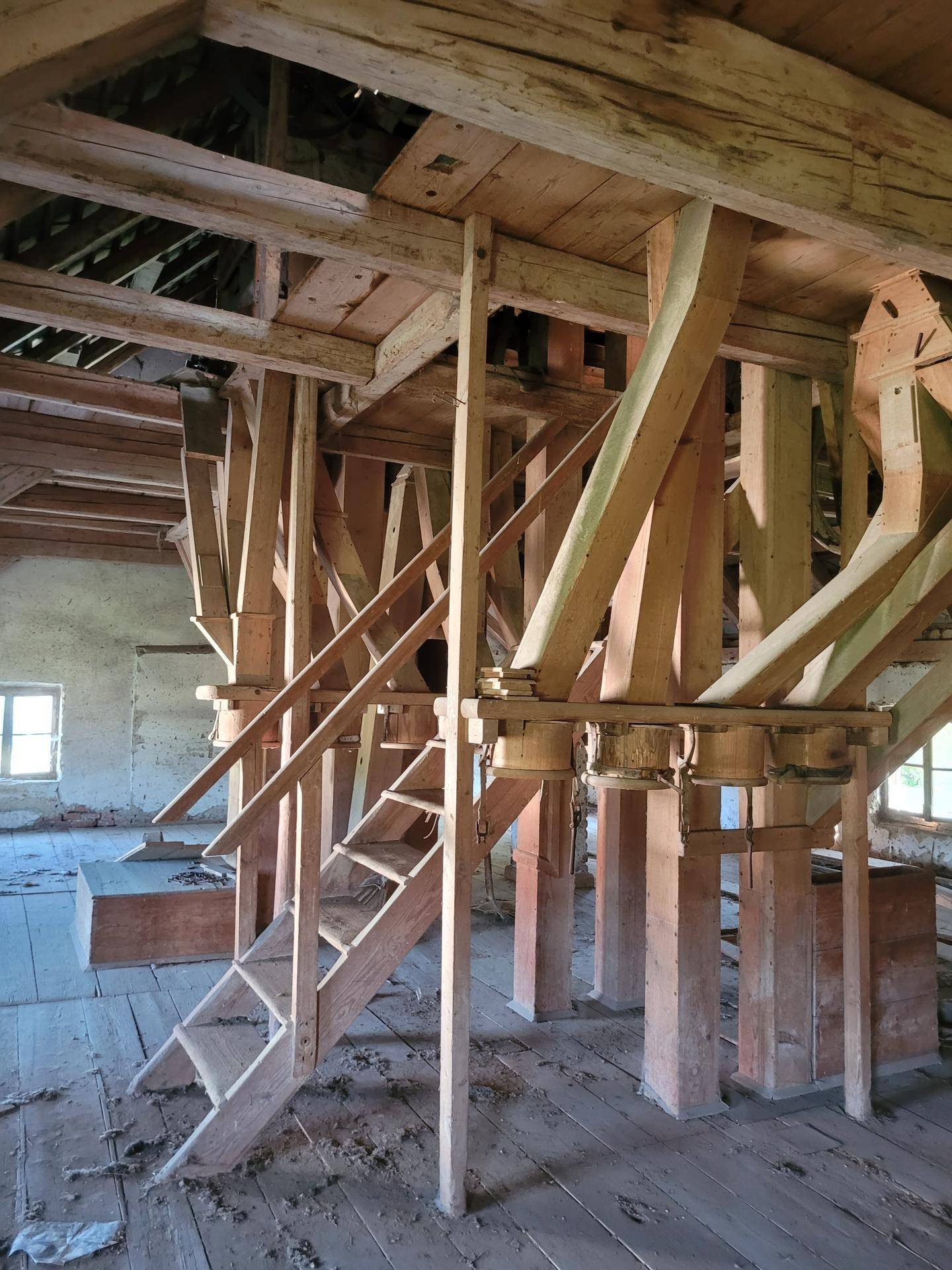 Innenansicht mit massiven Holzbalken, Holztreppe und Fenstern, die Tageslicht hereinlassen.