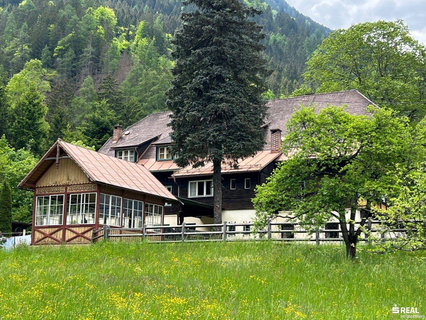 Traditionelles Gasthaus mit Anbau und weitläufiger grüner Wiese vor bewaldeten Bergen.