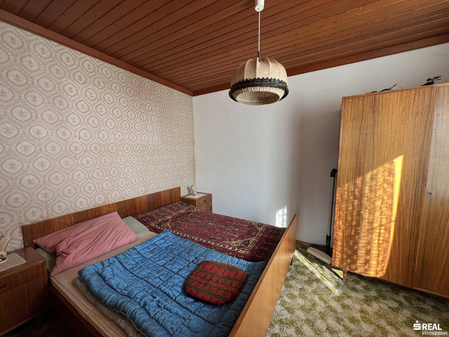 Schlafzimmer mit Holzdecke, Teppichboden und einfacher Einrichtung im Landhausstil.