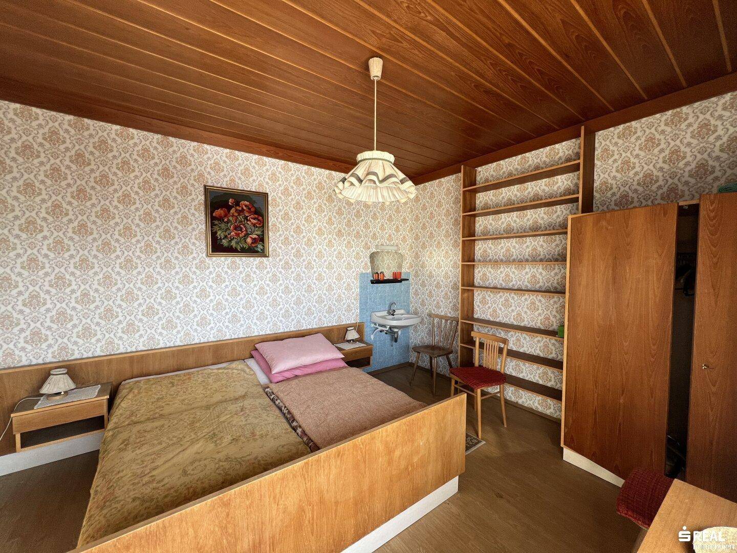 Schlafzimmer mit Holzdecke, gemusterter Tapete, Holzregalen und Waschbecken.